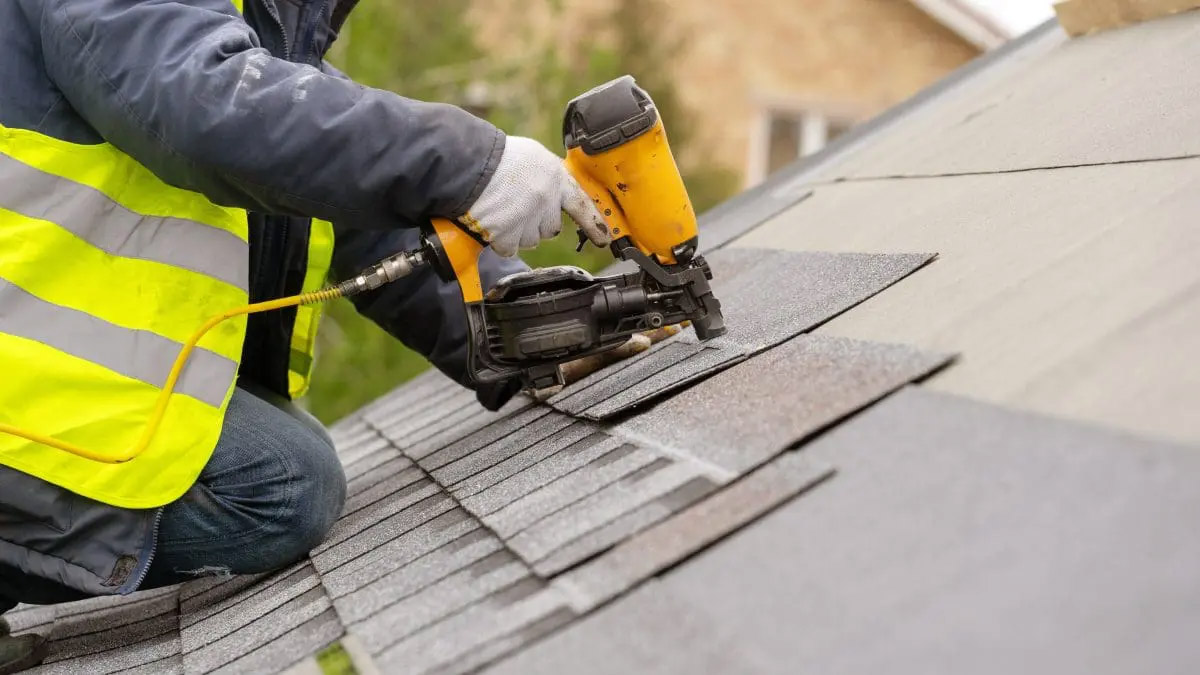 A close-up of a roofer wearing PPE and using a nail gun to install a shingle roof