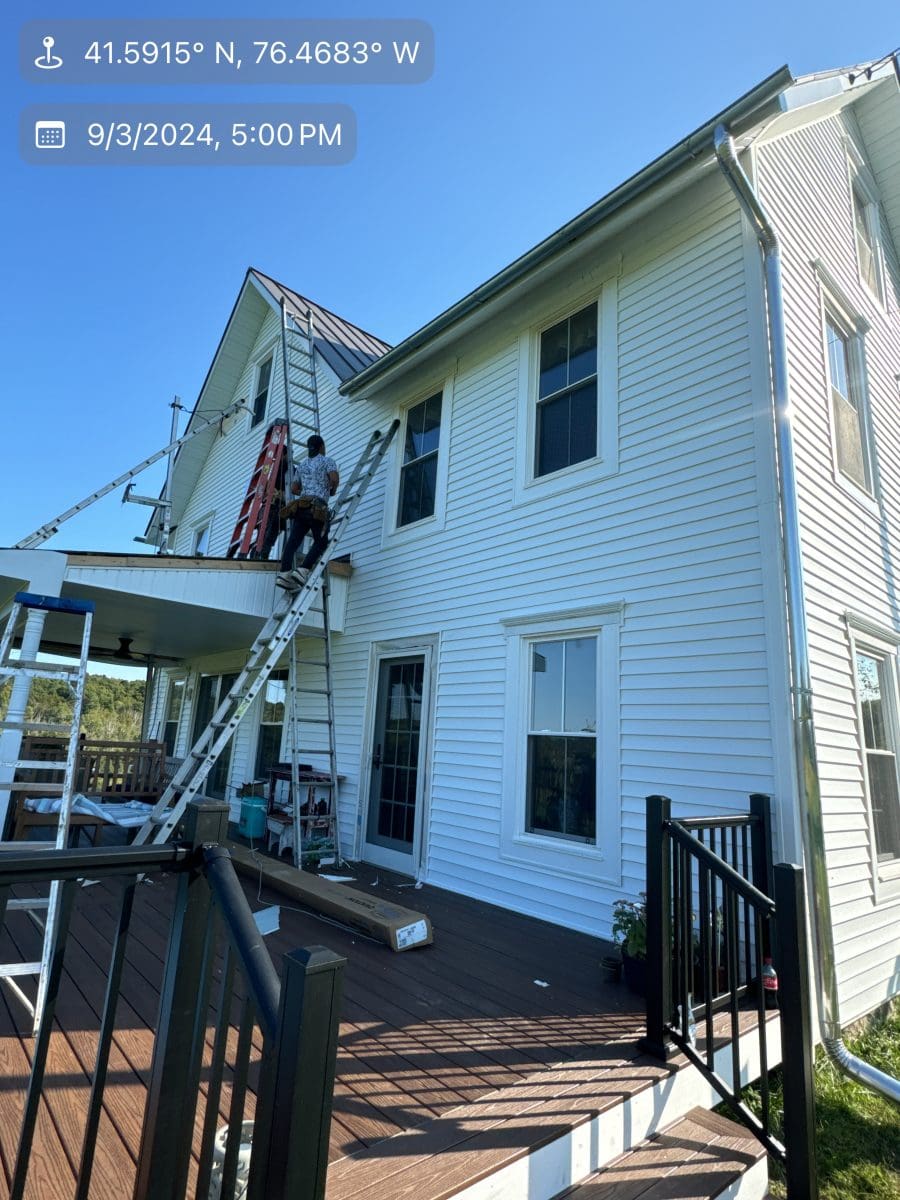 A white two-story home with ladders leaning against it as workers replace siding.