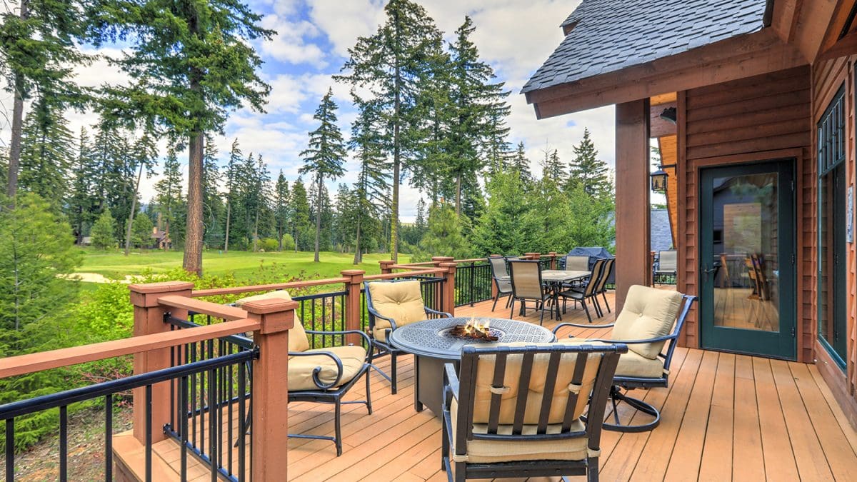 A large wooden cabin home to the right, with the focus of the image on a large wooden deck that holds two tables with chairs around them.