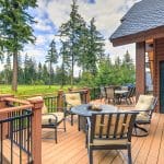 A large wooden cabin home to the right, with the focus of the image on a large wooden deck that holds two tables with chairs around them.