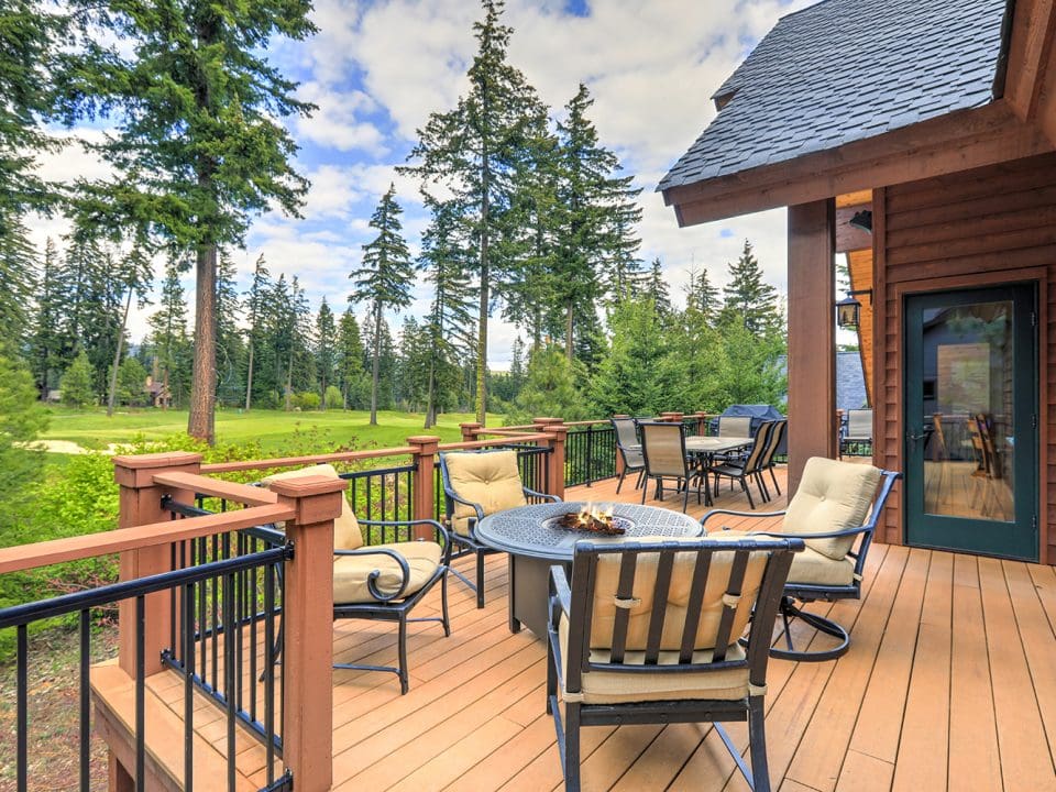 A large wooden cabin home to the right, with the focus of the image on a large wooden deck that holds two tables with chairs around them.