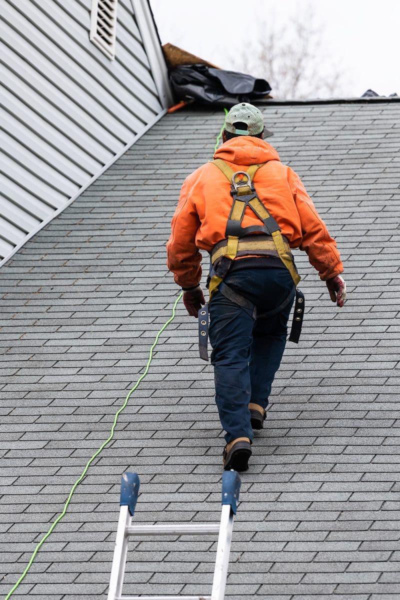 A roofing contractor wearing safety gear walks up a roof